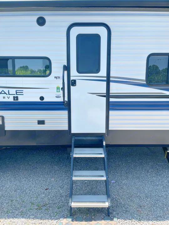 A white and blue RV rental travel trailer stands ready with its steps deployed on a gravel campsite, inviting adventure.. Keystone RV Springdale 2023