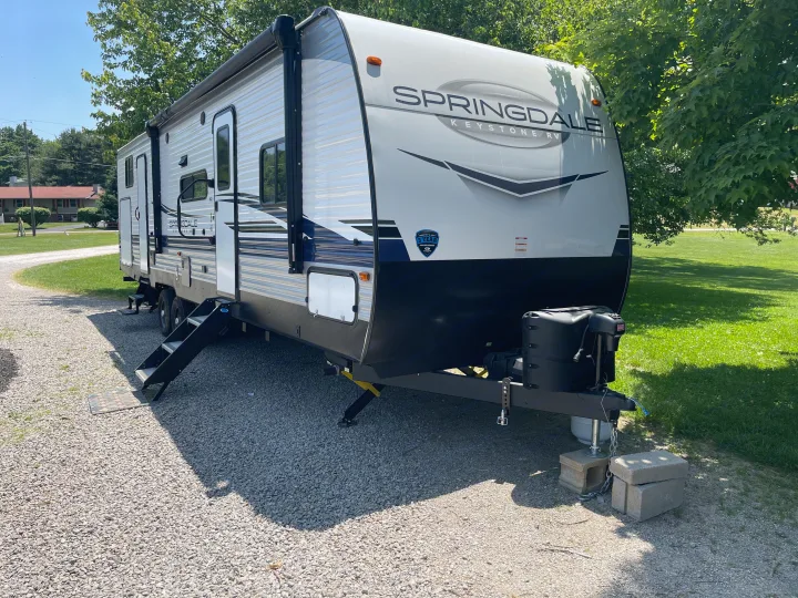 A white Springdale Keystone travel trailer RV, ready for your next rv rental, parked on a sunny gravel lot with green trees.. Keystone RV Springdale 2023