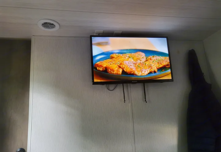 Wood-paneled RV rental interior with a TV displaying cooked food. A ceiling vent and hanging jacket are visible.. Forest River Wildwood X-Lite 2022