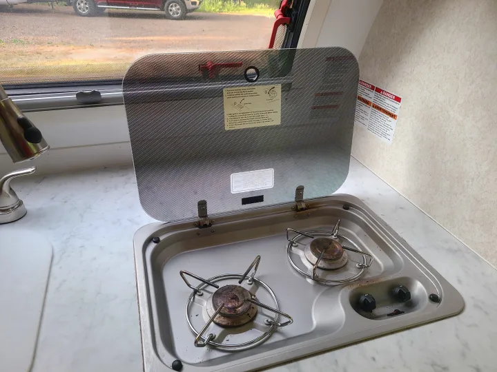 RV rental kitchen: a two-burner gas stove with its glass cover open, showing instructions and warning labels for cooking.. Xtreme Outdoors Little Guy 2019