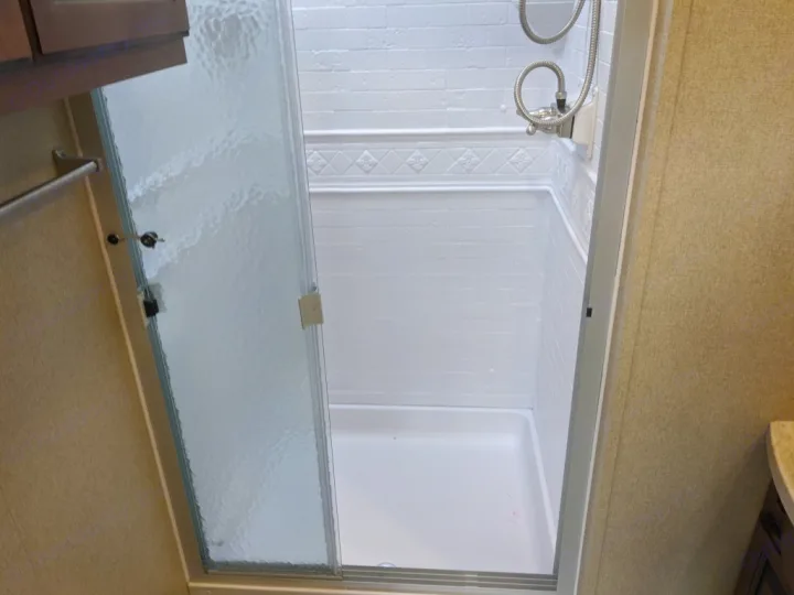 Bright, compact RV rental shower with white tiled walls, shower head, and clear glass sliding door.. Grand Design Reflection 2018
