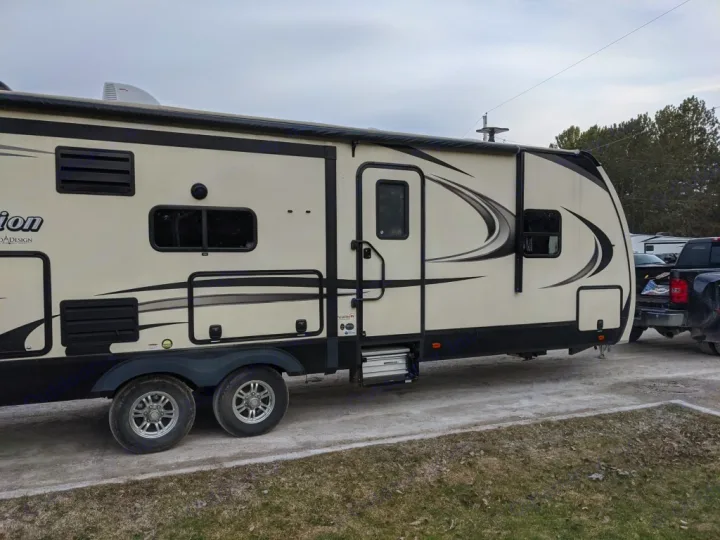 Side view of a large, light-colored travel trailer, an RV rental, hooked to a truck on a gravel lot. Perfect for camping trips.. Grand Design Reflection 2018
