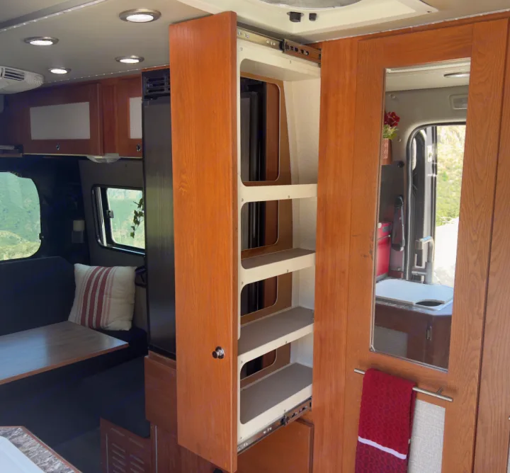 Inside an RV rental: a pull-out pantry with shelves, mirrored door with towel bar, and a dining area by a window.. Roadtrek Simplicity SRT 2019