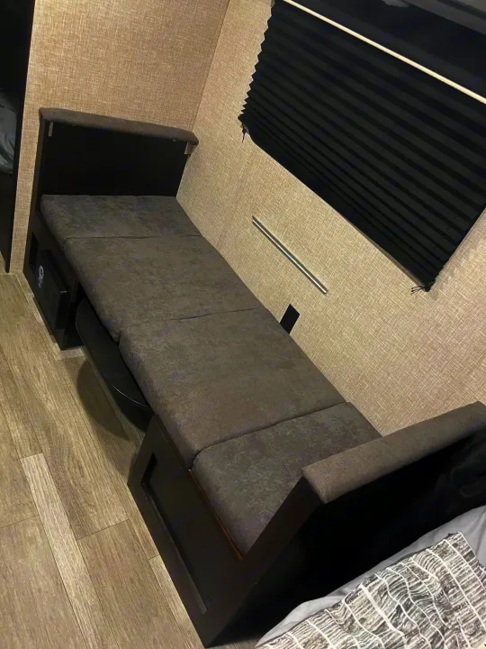 Inside an RV rental: A compact gray sofa converts to a bed, featuring a pull-out, by a window with a black blind.. Jayco Jay Flight SLX 2023