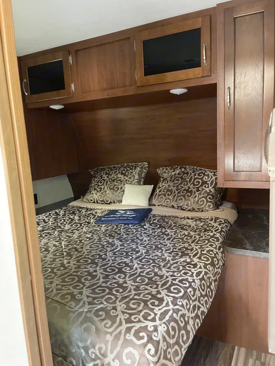 Inside an RV rental: a compact bedroom with a patterned bed, overhead cabinets, and built-in storage for travel comfort.. Jayco Jay Flight 2017