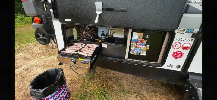 This RV rental's outdoor kitchen features a griddle cooking bacon, a sink, and a mini-fridge, ideal for camping meals.. Winnebago Minnie 2022