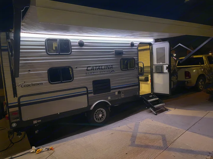This RV rental is a grey Coachmen Catalina travel trailer, illuminated at night with its awning lights and open door.. Coachmen Catalina Summit 2022
