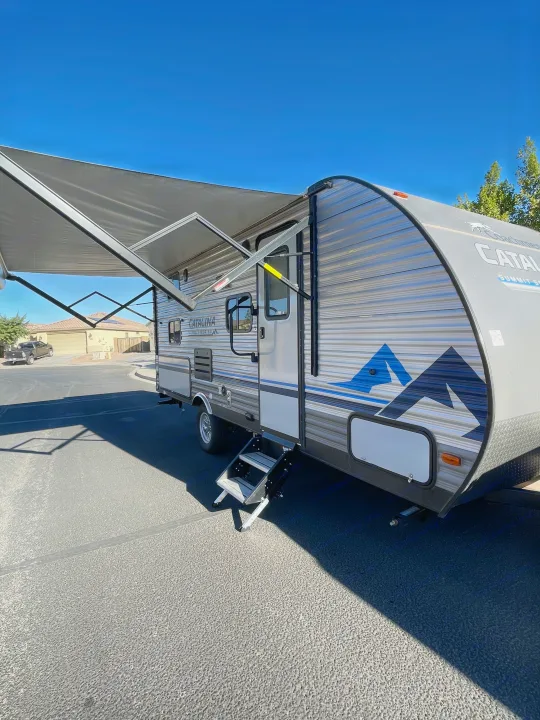 A grey Catalina travel trailer (RV rental) with its awning extended and steps down, parked on asphalt on a sunny day.. Coachmen Catalina Summit 2022