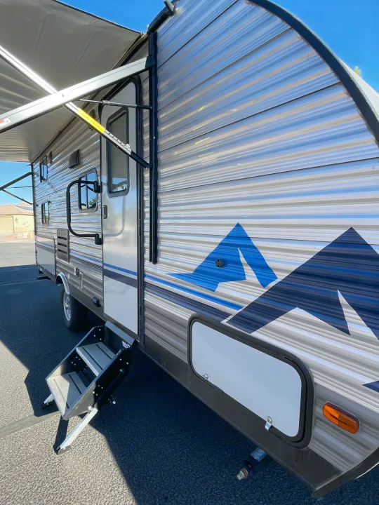 Side view of an RV rental with awning extended and steps deployed, featuring a blue mountain graphic on its side.. Coachmen Catalina Summit 2022