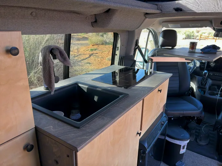 RV rental: Inside a camper van, a compact kitchen with a sink, induction cooktop, and front swivel table. View of desert landscape.. Ford Custom Transit 2022