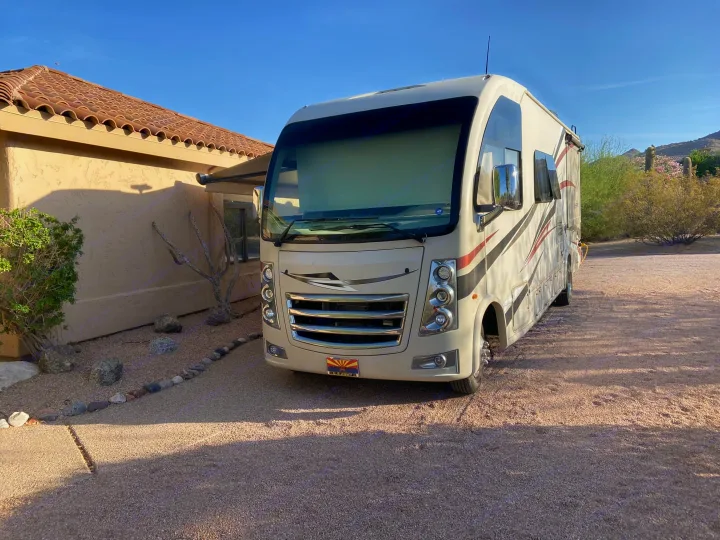 This large, beige motorhome, available for RV rental, is parked on a sunny gravel driveway beside a desert home.. Thor Vegas 2018