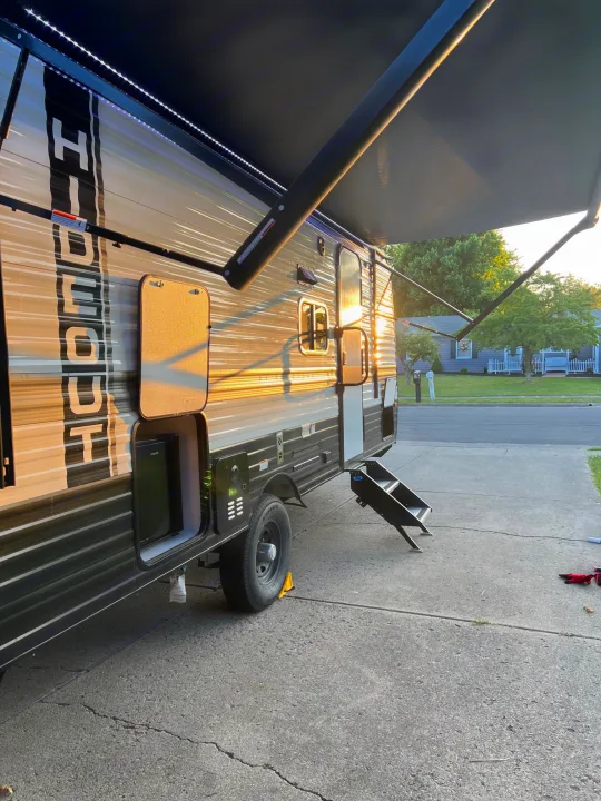 This rv rental is parked in a driveway with its awning extended, illuminated by LED lights and golden hour sun.. Hideout 181BH 2023