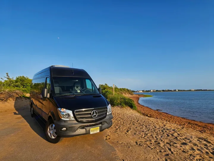 A black Mercedes Sprinter van, likely an RV rental, parked on a dirt path next to a serene bay under a clear sky.. Mercedes-Benz Sprinter 2016