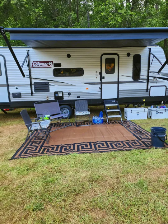Relaxing RV rental setup with extended awning, outdoor rug, and patio furniture on grass next to trees.. Coachmen lantern Dutchman 263 BHWE 2021