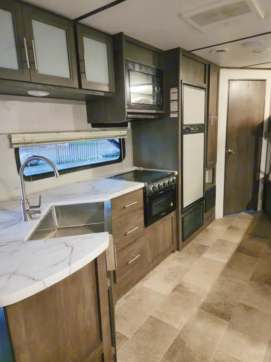 Explore this RV rental kitchen's sleek design: microwave, fridge, stove/oven, and a large sink, all with dark wood cabinetry and tiled floors.. Coachmen lantern Dutchman 263 BHWE 2021