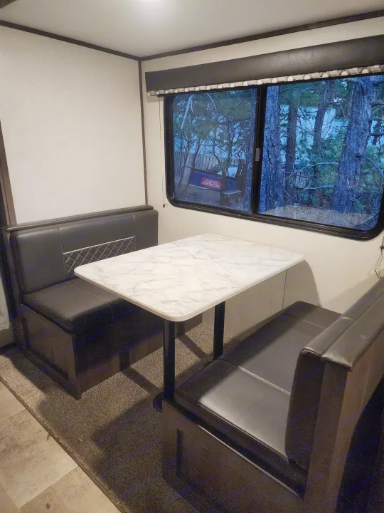 Inside an RV rental: Dinette area with a white marble table, black benches, and a large window looking out at trees.. Coachmen lantern Dutchman 263 BHWE 2021