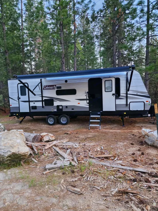 A modern travel trailer, ready for your RV rental adventure, parked at a rustic forest campsite surrounded by tall pines.. Coachmen lantern Dutchman 263 BHWE 2021