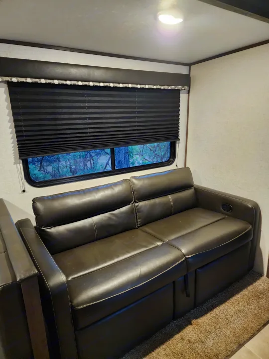RV rental interior: A dark leather sectional sofa beside a window with pleated blinds, showing trees outside, and a wall light.. Coachmen lantern Dutchman 263 BHWE 2021