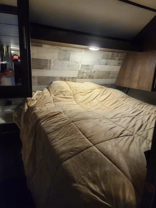A cozy RV rental bedroom features a large bed with a tan quilted comforter, wood-look floor, and the photographer's reflection.. Coachmen lantern Dutchman 263 BHWE 2021
