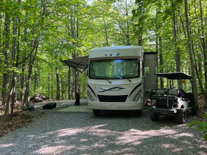 Enjoying nature with our RV rental! A beige motorhome with an awning extended, parked on gravel next to a golf cart, surrounded by lush green trees.. Thor Motor Coach A.C.E 2017