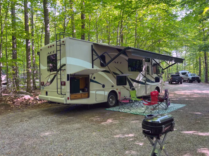 A beige RV rental is parked at a wooded campsite with an awning, chairs, and a grill, offering comfortable camping.. Thor Motor Coach A.C.E 2017