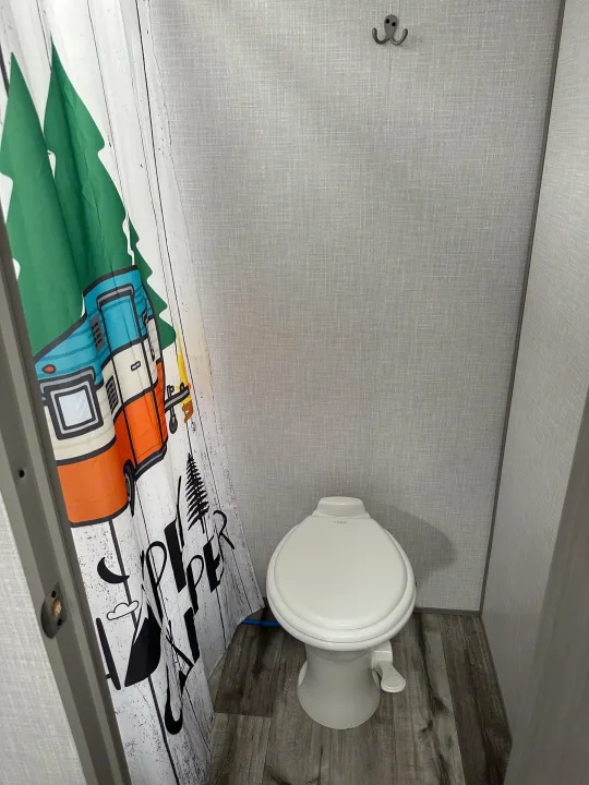 Compact RV rental bathroom features a white toilet, wood-look floor, and a fun camping-themed shower curtain. A hook is on the wall.. Dutchmen Coleman Lantern LT 2022