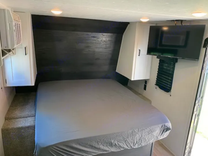 Cozy RV rental bedroom with grey bed, dark headboard, white cabinets, mounted TV, AC unit, and access steps.. Keystone RV Hideout 2021