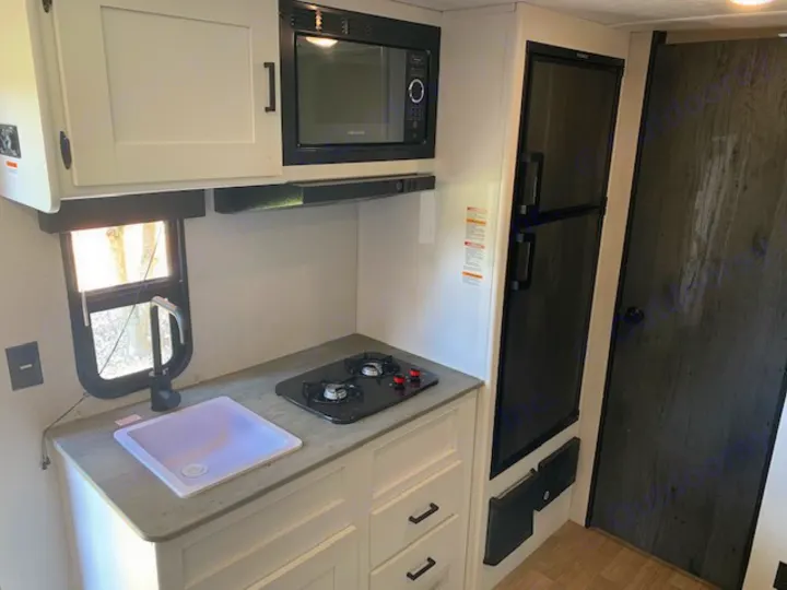 Modern RV rental kitchen with white cabinets, microwave, sink, 2-burner stove, and refrigerator.. Keystone RV Hideout 2021