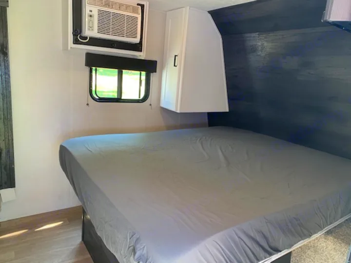 Cozy RV rental bedroom with gray bed, AC unit, and window. Overhead cabinet visible. Ready for your adventure.. Keystone RV Hideout 2021