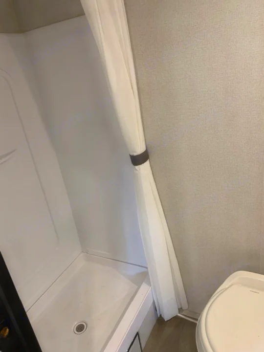 Explore an RV rental's compact bathroom: a bright white shower with curtain tied back, next to the toilet.. Keystone RV Hideout 2021
