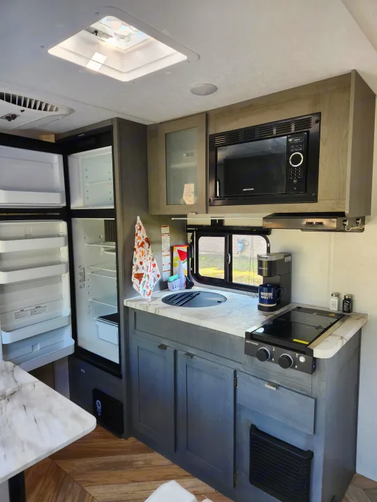 RV rental interior: A modern, compact kitchen featuring a microwave, stove, sink, and open refrigerator, ideal for travel.. Ozark Ozark Trailer 2023