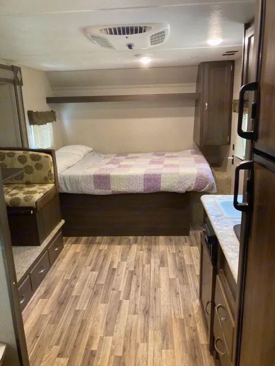 Interior view of an RV rental showing a compact bedroom with a purple quilt, a kitchen counter, and a small seating area.. Keystone RV Springdale 2019