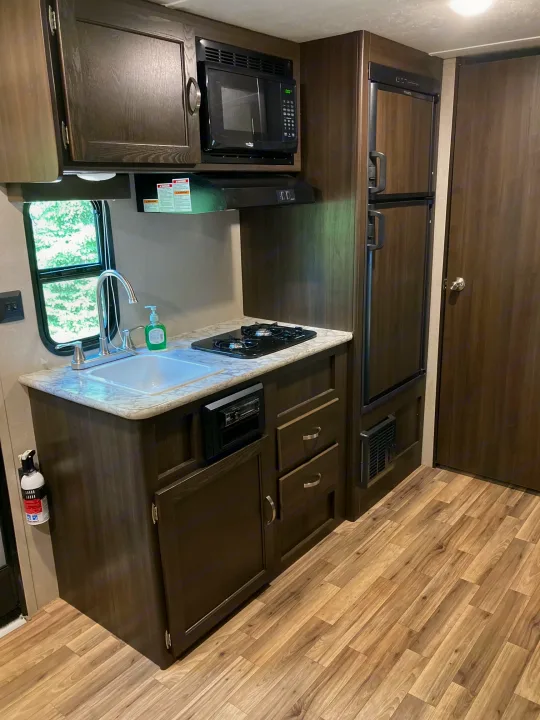 Inside an RV rental: full kitchen with dark cabinetry, microwave, stove, sink, and refrigerator. Light wood floor.. Keystone RV Springdale 2019
