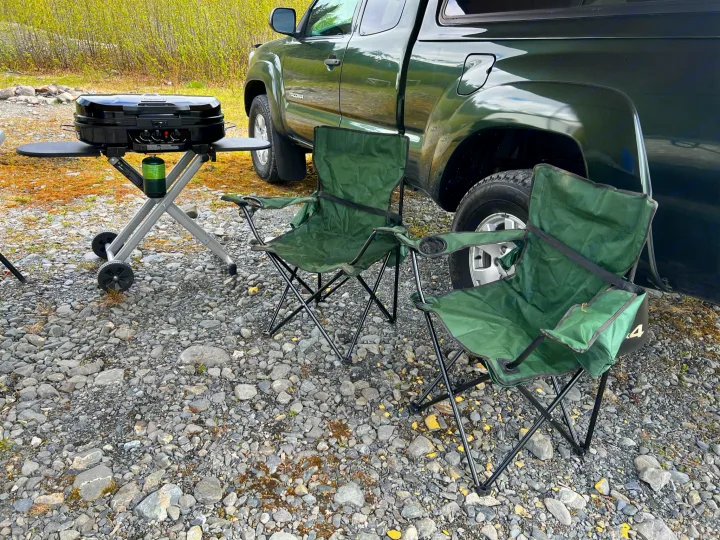 A green pickup truck, black portable grill with a propane tank, and two green camping chairs on gravel. Ideal for rv rental trips.. Toyota Tacoma 2012