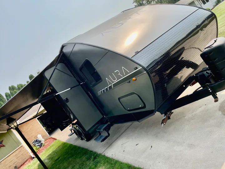 Dark gray Aura travel trailer, possibly an RV rental, parked with its awning extended and sun reflecting off its sleek front.. Travel Lite Aura 2019
