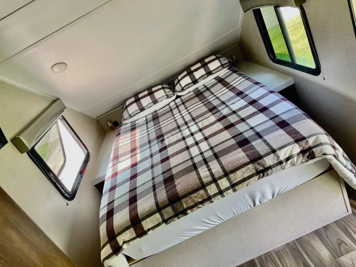 Serene RV rental bedroom featuring a comfy plaid bed, side windows showing greenery, and a ceiling light.. Travel Lite Aura 2019