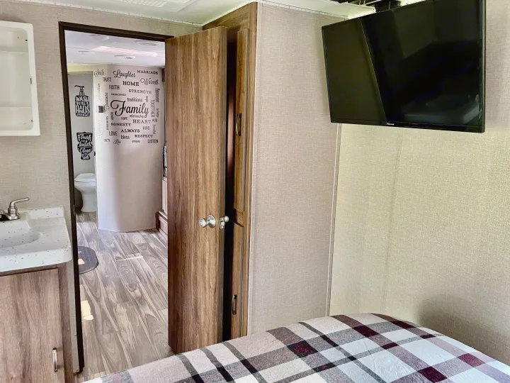 Explore this cozy RV rental interior: bedroom with TV, leading to a compact bathroom with sink, toilet, and fun word decals.. Travel Lite Aura 2019