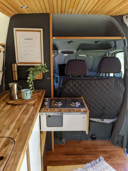 Explore this stylish RV rental: a compact camper van kitchen with wooden counters, stove, and Canmore-themed art.. Mercedes-Benz Mercedes Sprinter 2500 2015