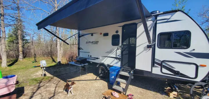 RV rental at a sunny campsite with an extended awning, outdoor grill, and playful dog by the travel trailer.. Forest River Surveyor Legend 2022