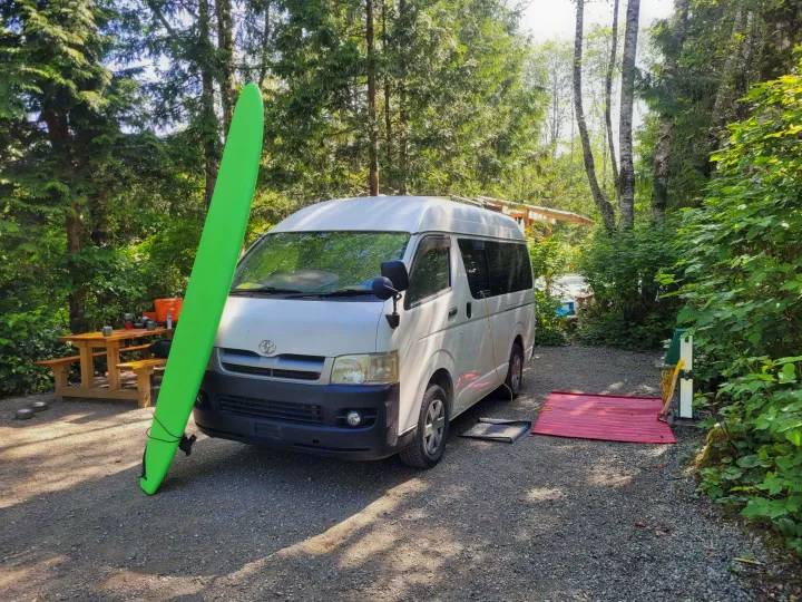 Enjoying the outdoors with an RV rental: a white camper van, surfboard, and picnic setup at a lush green campsite.. Toyota Hiace 2006