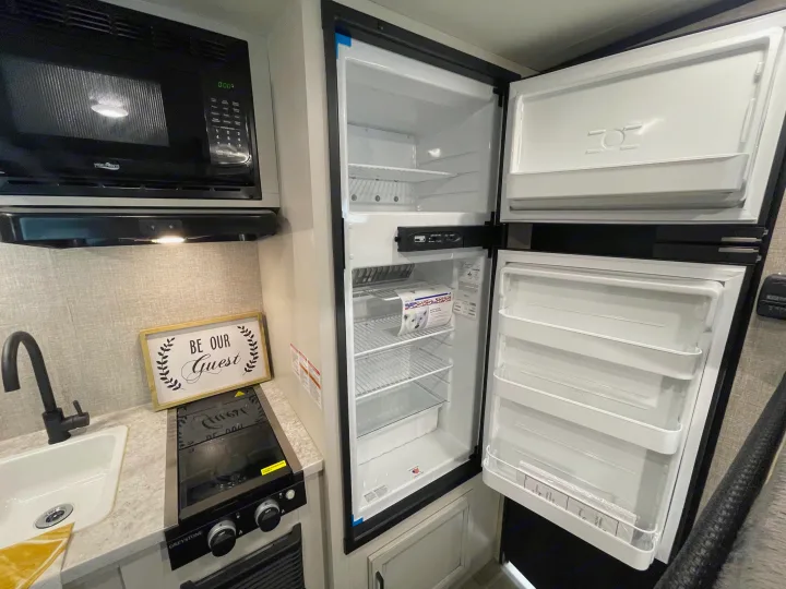 RV rental kitchenette: open fridge, microwave, stove, and sink. A "Be Our Guest" sign adds a welcoming touch.. Jayco Jay Flight 2022