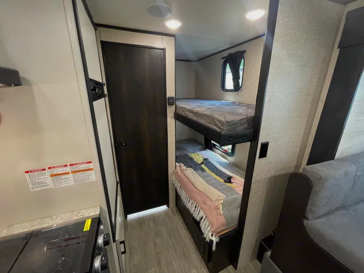 RV rental interior features compact bunk beds with an emergency exit window, a dark door, and a kitchen area, for comfortable travel.. Jayco Jay Flight 2022