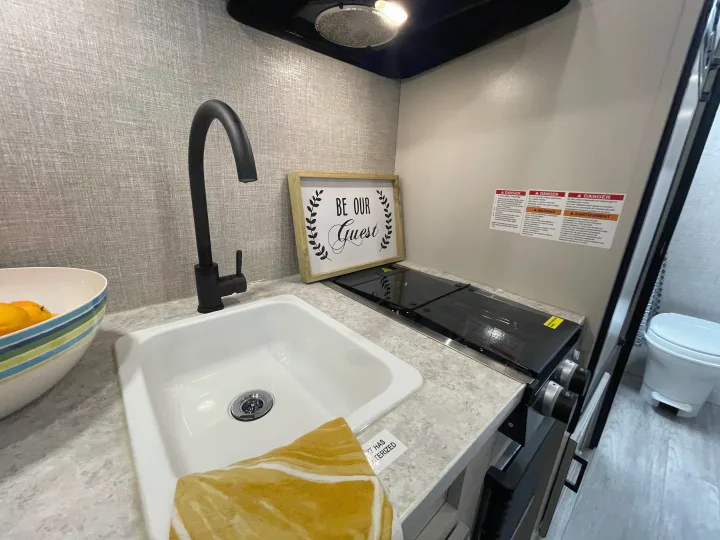 An RV rental's compact galley kitchen features a sink, black faucet, cooktop, and a welcoming "Be Our Guest" sign.. Jayco Jay Flight 2022