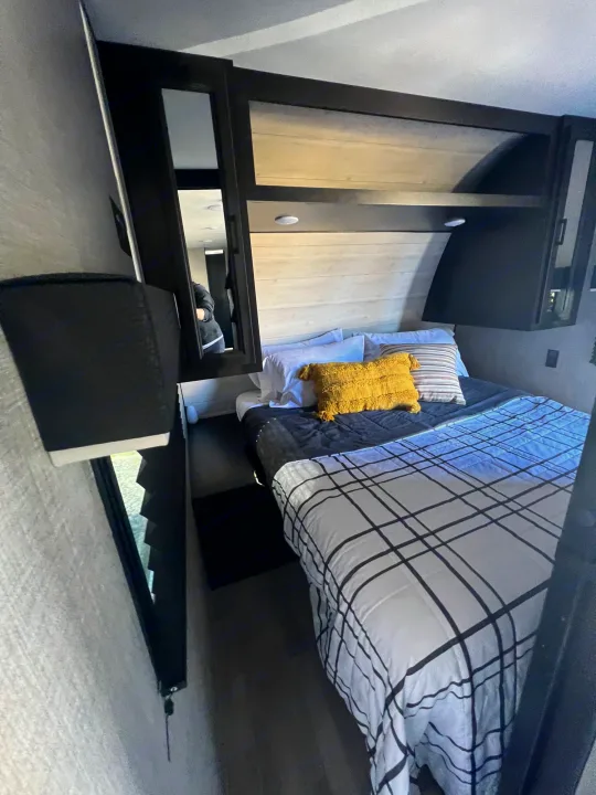 This comfortable RV rental bedroom features plaid bedding, dark overhead cabinets, and a mirror reflecting the interior.. Jayco Jay Flight 267BH 2023