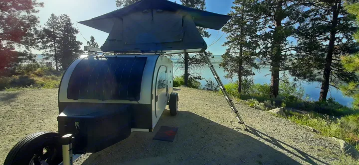 Lakeside RV rental: Teardrop trailer with deployed rooftop tent among pine trees on a sunny day, perfect for camping.. droplet XL 2026