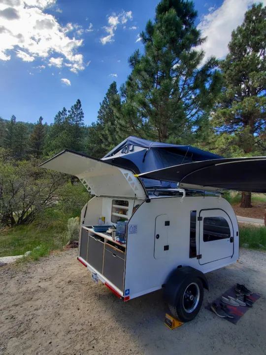 A white teardrop RV rental with an open rear kitchen and a rooftop tent is parked at a scenic, wooded campsite.. droplet XL 2026