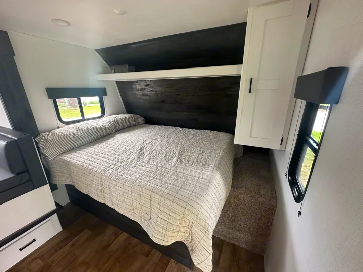 Inside your RV rental: a modern, compact bedroom with a cozy bed, striped pillow, dark wood headboard, and windows.. Keystone RV Hideout 2023
