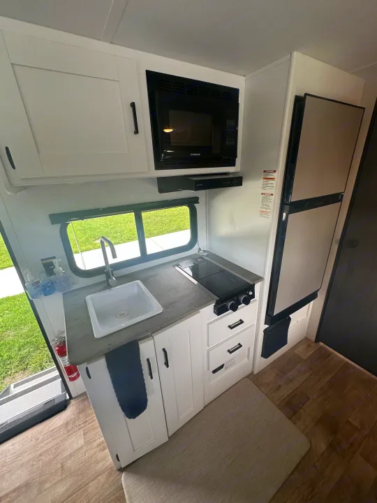This RV rental kitchen features white cabinets, sink, stove, microwave, and fridge, showcasing a compact and functional layout.. Keystone RV Hideout 2023