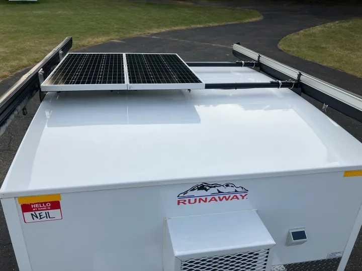 This white Runaway camper, perfect for RV rental, features rooftop solar panels & a side awning. A "Hello, my name is Neil" sticker is on the front.. Runaway Campers Rangerunner 2021