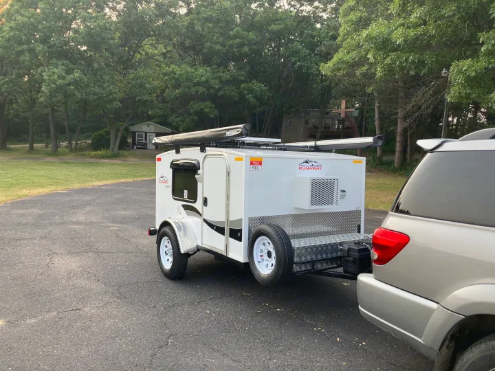 Runaway RV rental camper hitched to an SUV, parked on asphalt in a wooded campground. Perfect for camping adventures!. Runaway Campers Rangerunner 2021